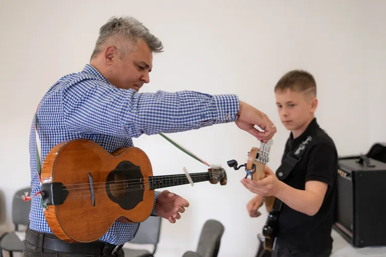 A zenekarvezető egy fiú hangszerének fejét igazítja, miközben mindketten tamburát tartanak a próbateremben.