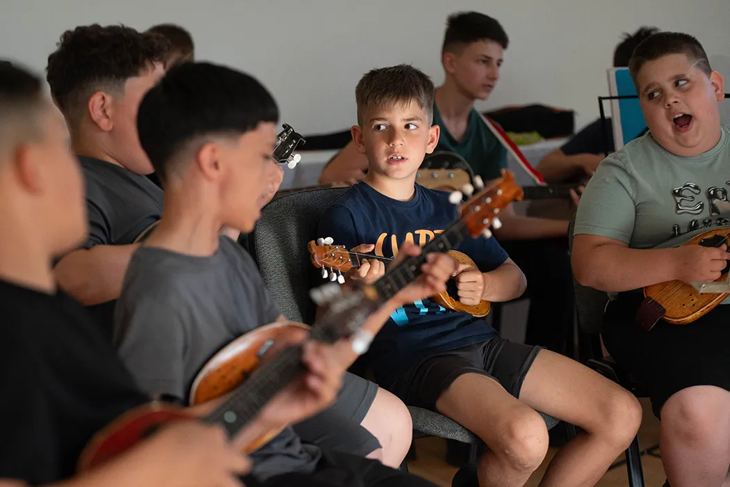 Tamburán játszó fiúk ülnek egymás mellett a próbateremben, középen egy sötétkék pólós gyerek figyel.