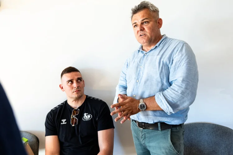 Zenekarvezető beszél, mellette ülő PICK Szeged játékos figyel a sükösdi teremben