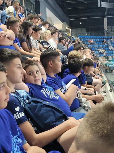 Kék pólós gyerekek egymás mellett a PICK Aréna lelátóján, és a kézilabda-bajnokság döntőjét nézik.