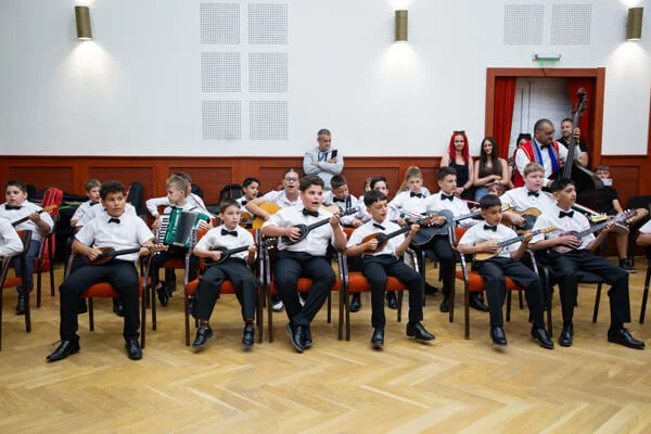 A SUGO Tamburazenekar gyermekei egy kupacban ülve játszanak tamburán és harmonikán a közös próbán.