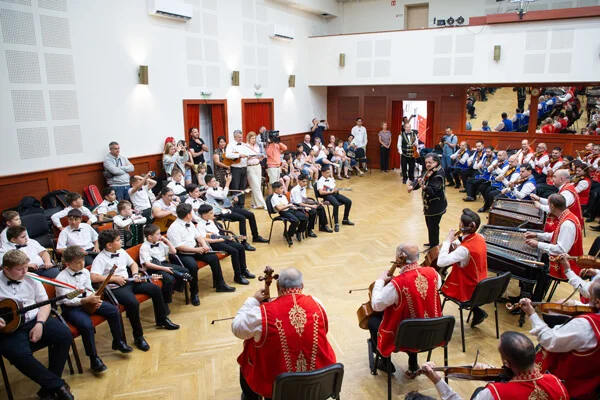 A két zenekar felülnézetben, egymással szemben ül a teremben, középen egy fekete ruhás nő áll.