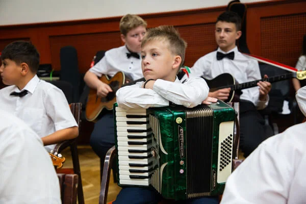 Fiú összefont karral egy zöld harmonikára támaszkodik, mögötte két társa gitárral ül a próbateremben.