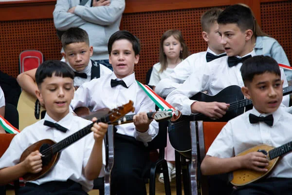 Hat fiú tamburán és gitáron játszik, a középen ülő gyerek magyar színű pánttal énekel a próbán.
