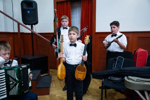Fiú két különböző tamburát tart a kezében, mellette harmonikás ül, mögötte társai hangszerekkel állnak.