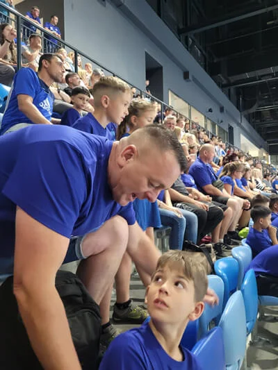 Egy felnőtt beszélget egy kisfiúval a lelátón, miközben körülöttük a döntőre érkezett szurkolók ülnek.