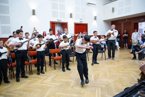 Egy csokornyakkendős fiú táncol a terem közepén, miközben mögötte a gyermek tamburások állva zenélnek.