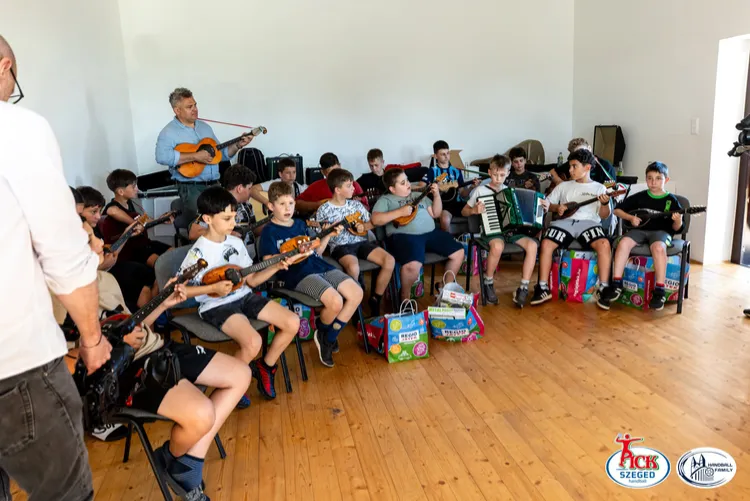 Az egész zenekar látszik, tamburán és harmonikán játszanak, mögöttük a zenekarvezető gitározik