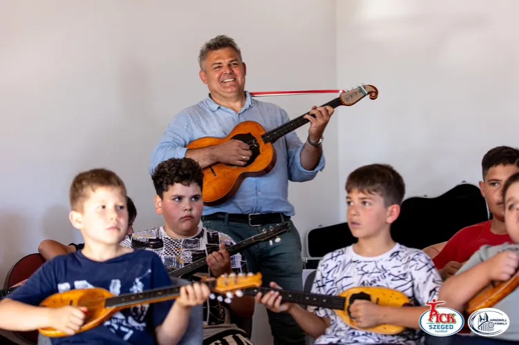 Zenekarvezető gitározik, miközben előtte ülő gyerekek tamburán játszanak