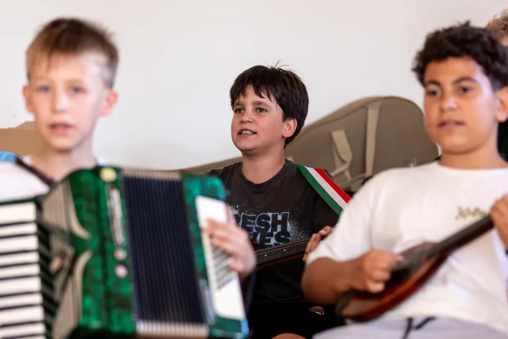 Harmonikán és tamburán játszó gyerekek zenélnek együtt, középen egy éneklő fiúval a próbateremben