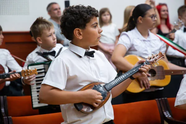 Göndör hajú fiú kis tamburán játszik, mögötte egy harmonikás és egy gitározó lány látható.