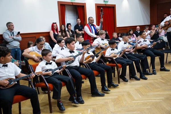 A SUGO Tamburazenekar tagjai egy hosszú sorban ülve játszanak, mögöttük felnőttek és egy nagybőgős áll.