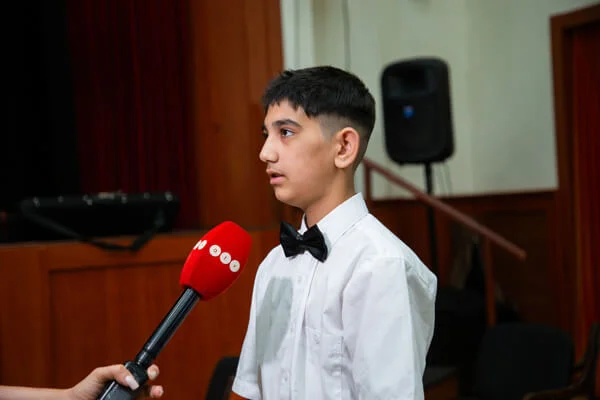 Fehér inges fiú oldalról látható, miközben egy piros mikrofont tartanak elé a rendezvényteremben.