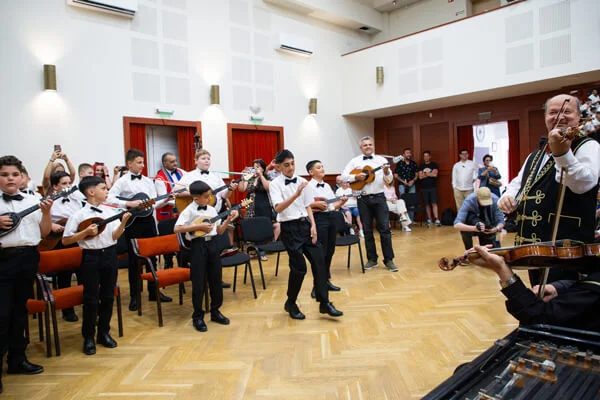 Tamburás gyerekek állnak félkörben, előttük egy fiú táncol, jobb oldalon hegedűs és cimbalom látható.