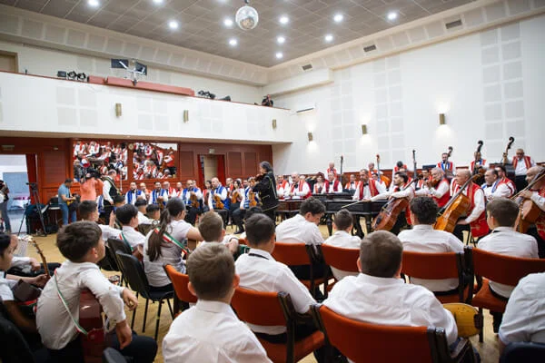 A gyermek tamburások hátulról láthatók, velük szemben a 100 Tagú Cigányzenekar nagybőgőkkel muzsikál.