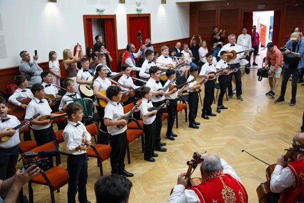 A gyermek tamburások állva, hosszú sorban zenélnek, előttük két piros mellényes felnőtt muzsikus ül.