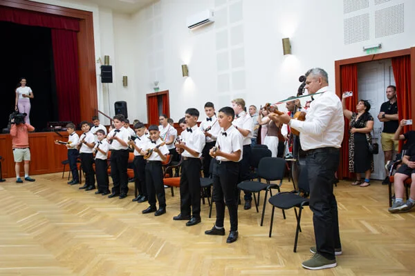 Fehér inges gyerekek egy sorban tamburáznak, jobb oldalon egy felnőtt zenész nagyméretű hangszeren játszik.