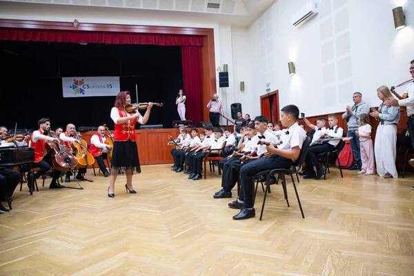 Hegedűs nő játszik a terem közepén, két oldalán a felnőtt zenekar és a gyermek tamburások ülnek.