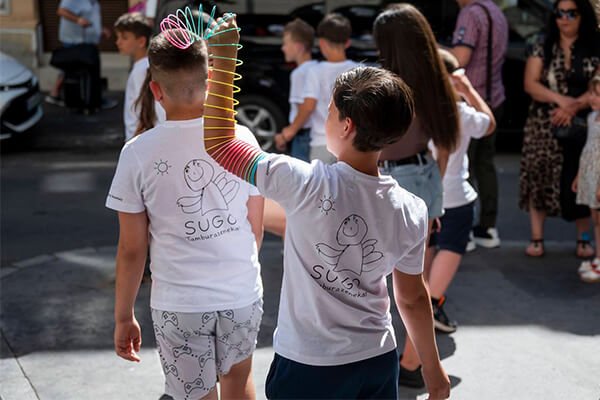 Két fiú fehér SUGO Tamburazenekar-pólóban áll a tömegben, egyikük szivárványszínű rugós játékkal.