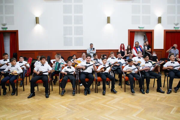 Csokornyakkendős gyerekek ülnek egy hosszú kupacban, tamburán, gitáron és harmonikán játszva a próbateremben.