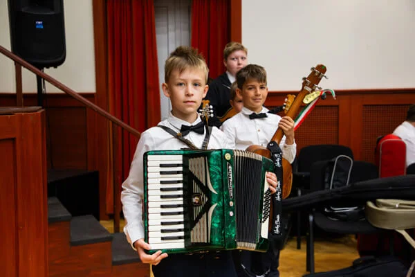 Egy fiú zöld harmonikát tartva néz a kamerába, mögötte társa hosszú nyakú tamburával és további gyerekek állnak.