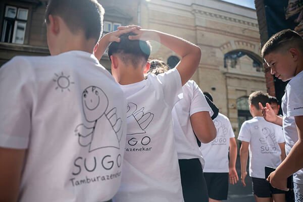 A SUGO Tamburazenekar fehér pólós gyermekei hátulról láthatók, egyikük a fejére teszi a kezét Budapesten.