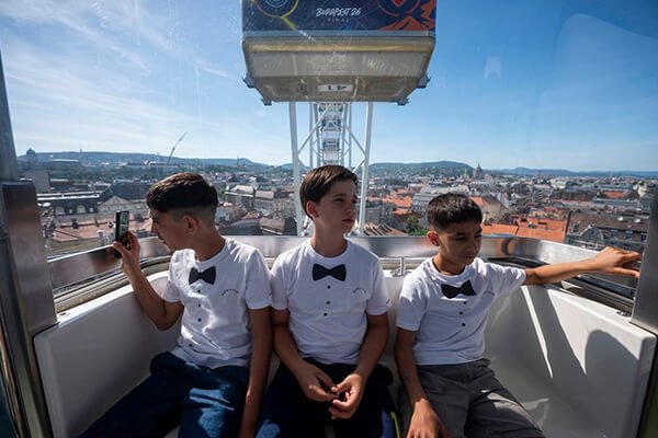 Három fiú csokornyakkendő-mintás fehér pólóban ül a Budapest Eye kabinjában, mögöttük a városi panoráma.
