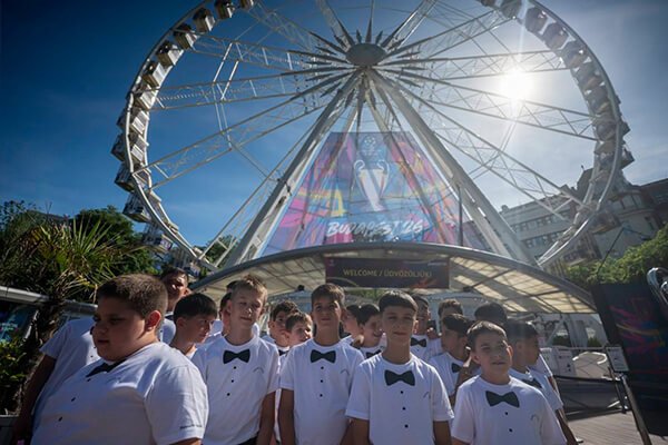 A SUGO Tamburazenekar gyermekei fehér, csokornyakkendős pólóban állnak csoportképen a Budapest Eye előtt