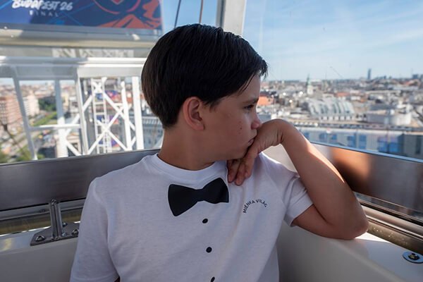 Fehér, csokornyakkendő-mintás pólót viselő fiú nézi Budapest panorámáját a Budapest Eye kabinjából.