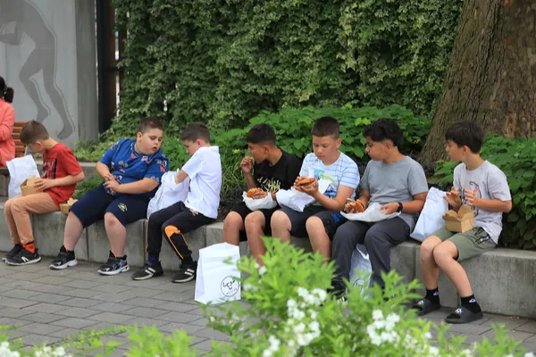 A Sugó Tamburazenekar gyerektagjai egy hosszú sorban ülve ebédelnek a Szent István park fái alatt.