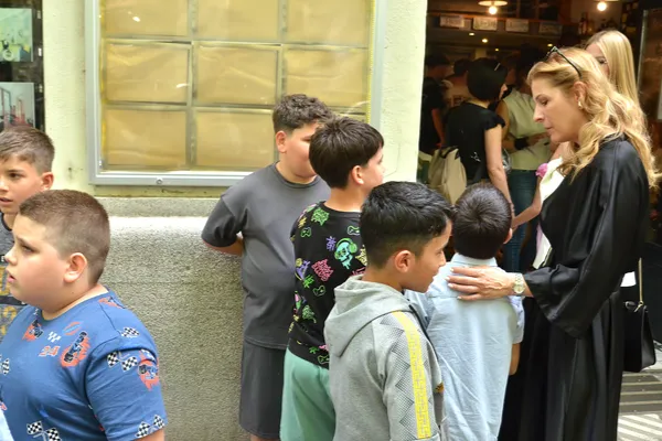 A zenekar gyerektagjai egy épület bejáratánál várakoznak, miközben egy nő egy kisfiúval beszélget.