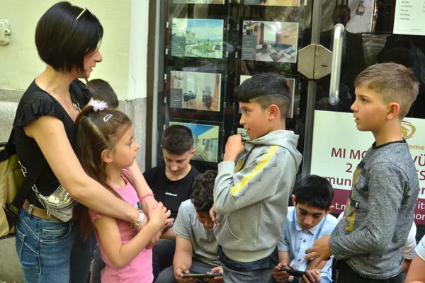 Egy nő egy kislányt ölelget, miközben a fiúk beszélgetnek és telefonoznak egy budapesti üzlet előtt.