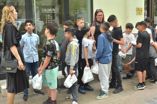 A zenekar gyerektagjai fehér csomagokkal gyülekeznek az épület előtt, kísérőik társaságában.