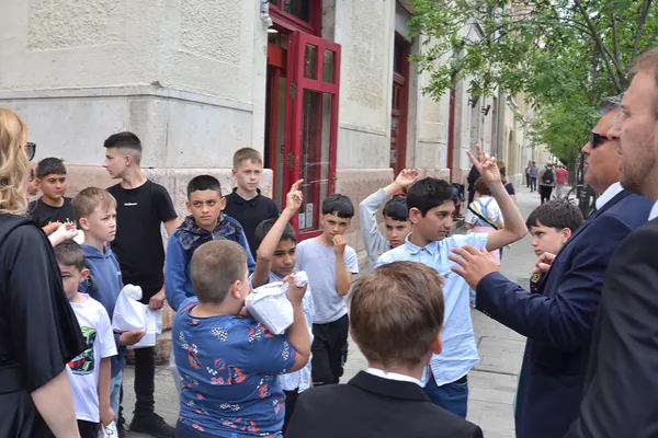 A gyerekek a zenekar vezetőjét hallgatják az utcán, miközben néhányan felemelik a kezüket, jelentkeznek.