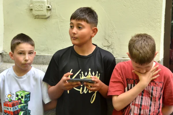 Három fiú áll az épület falánál, a középső telefont tart, társa pedig az arcához emeli a kezét.